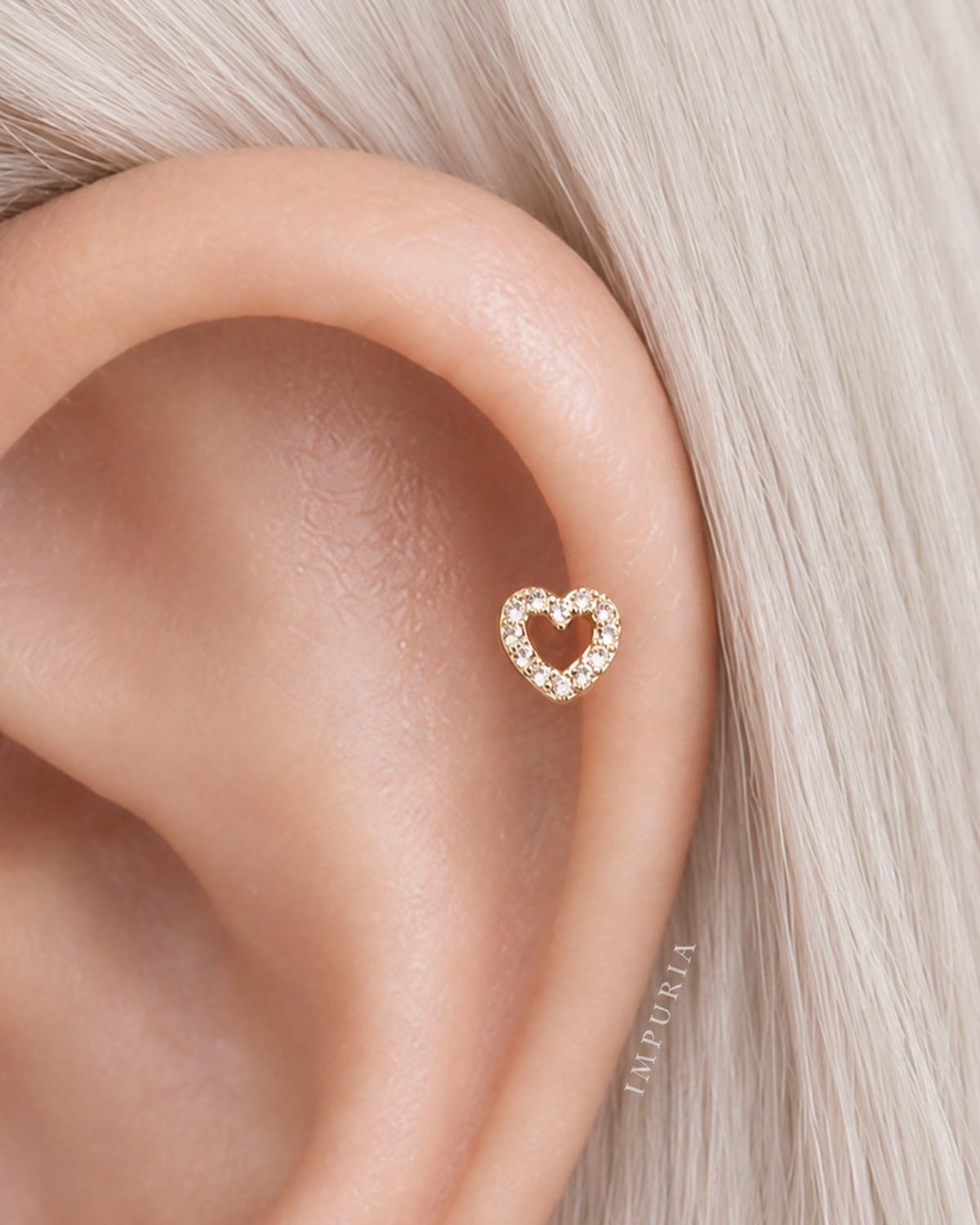 A close-up of a person's ear wearing a small gold heart-shaped stud earring with tiny clear stones, shown against pale blonde hair.