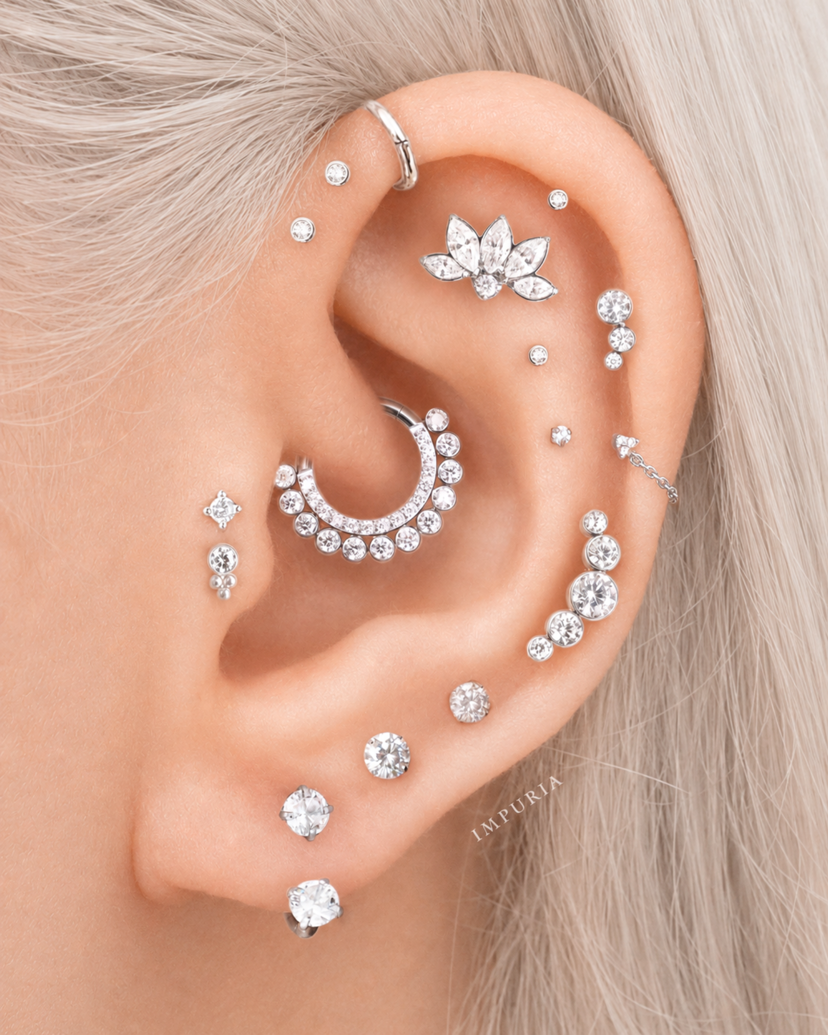 A close-up of a person's ear adorned with multiple silver-toned cartilage and lobe piercings, including hoop, stud, and floral-shaped earrings set with clear stones. The person has light blonde hair, and a small 'IMPURIA' brand watermark is visible on the earlobe.