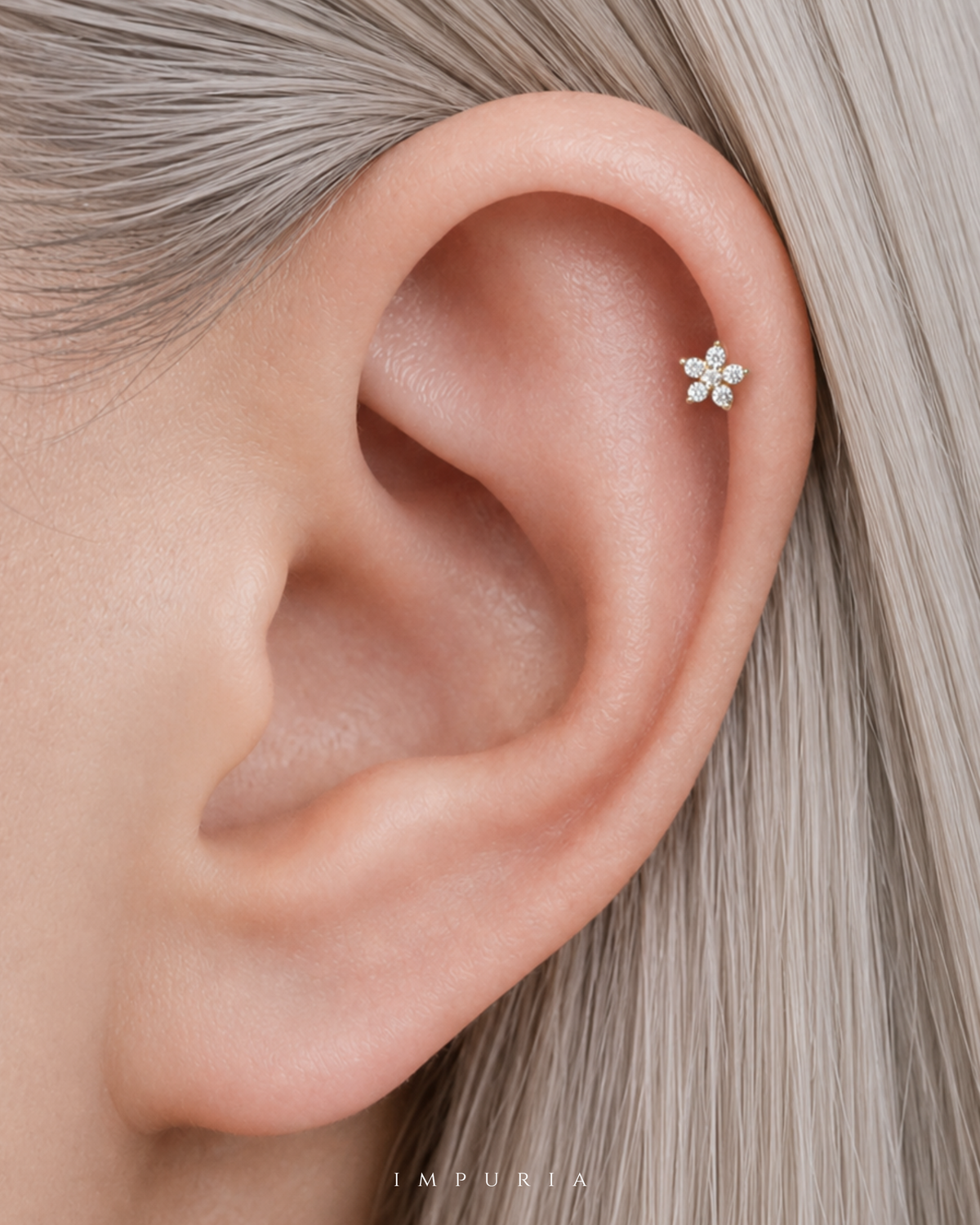 A close-up of a person's ear wearing a small silver flower-shaped stud earring with clear stones. The person has light silver-gray hair, and the brand name 'IMPURIA' is printed at the bottom.