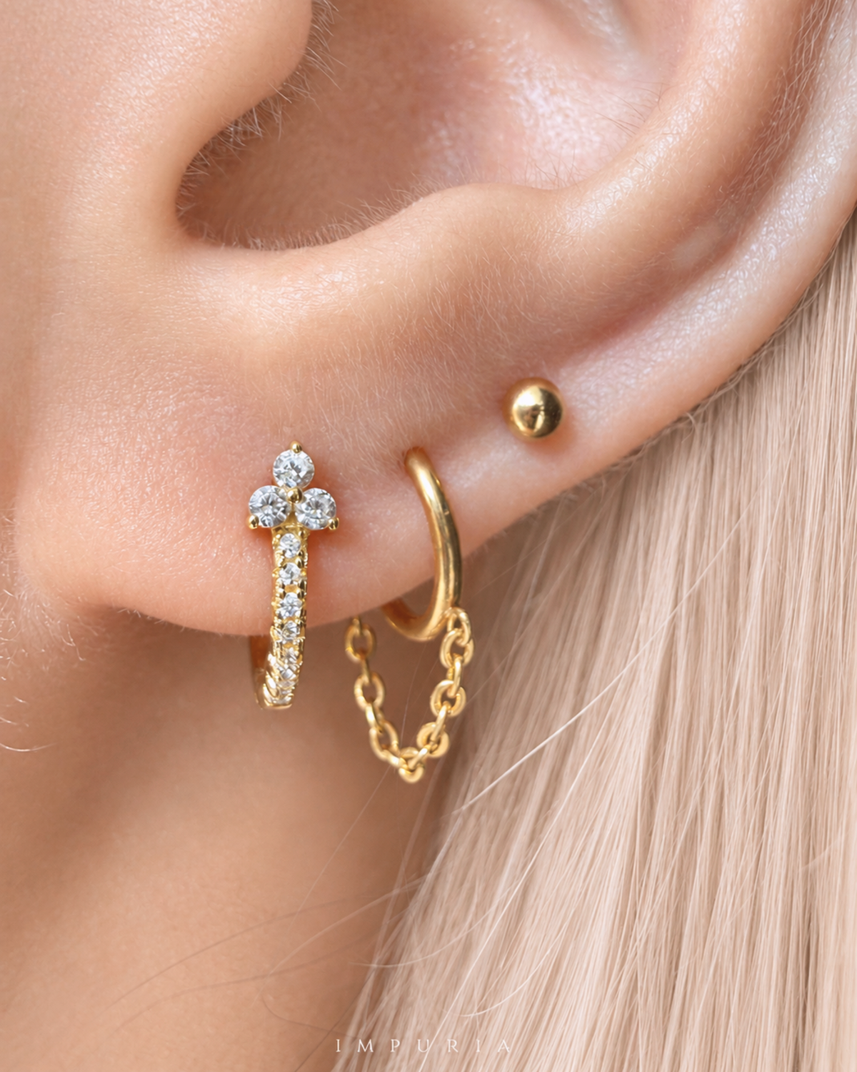 A close-up of a person's ear showcasing a gold hoop earring with a delicate chain drop and a small gold cross stud adorned with clear crystals, alongside a simple gold stud on the earlobe.
