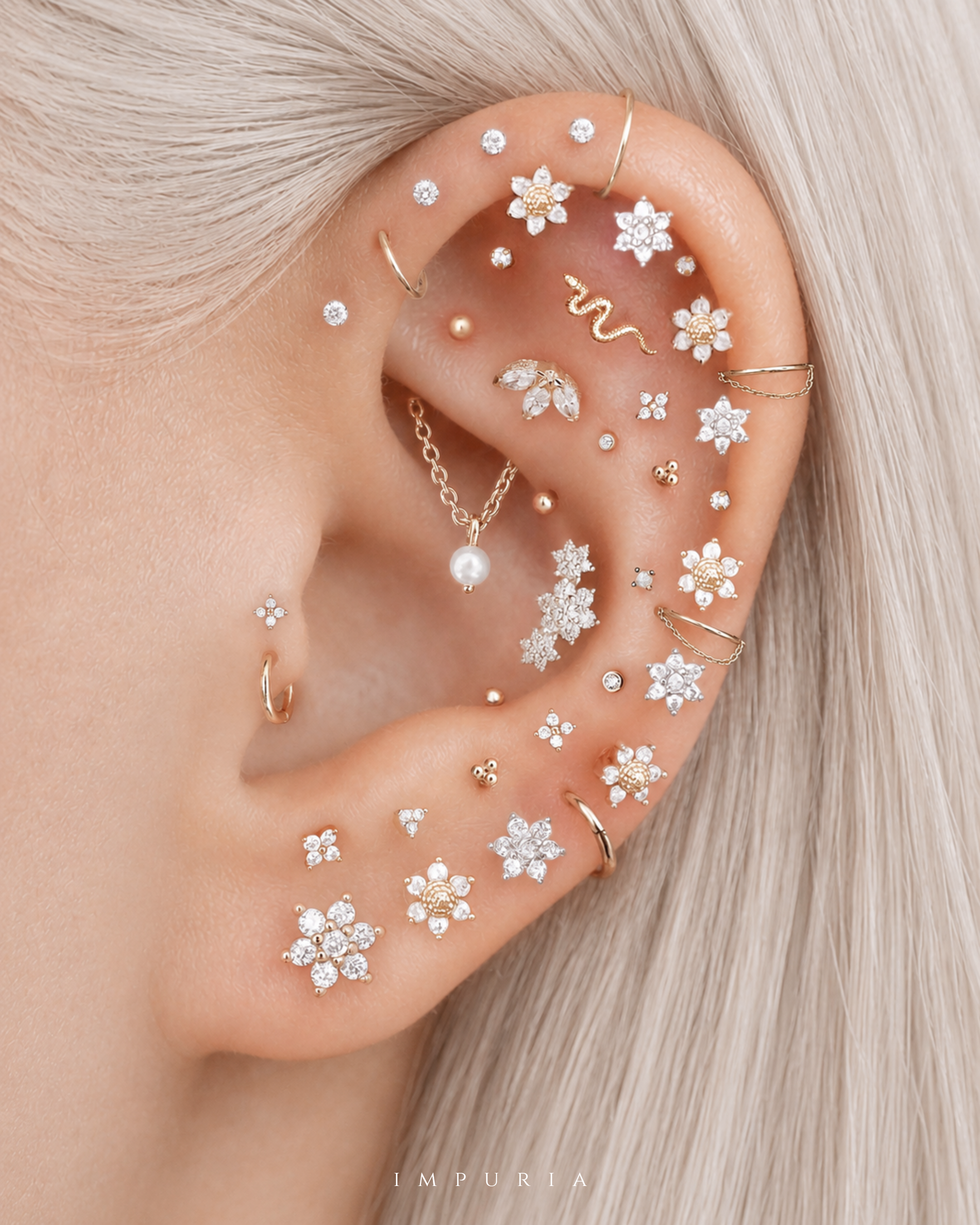 A close-up of a person's ear adorned with multiple delicate gold and silver ear piercings, including flower-shaped studs, hoop earrings, a small chain with a pearl drop, and ear cuffs, set against light blonde hair. The brand name 'IMPURIA' is visible at the bottom.
