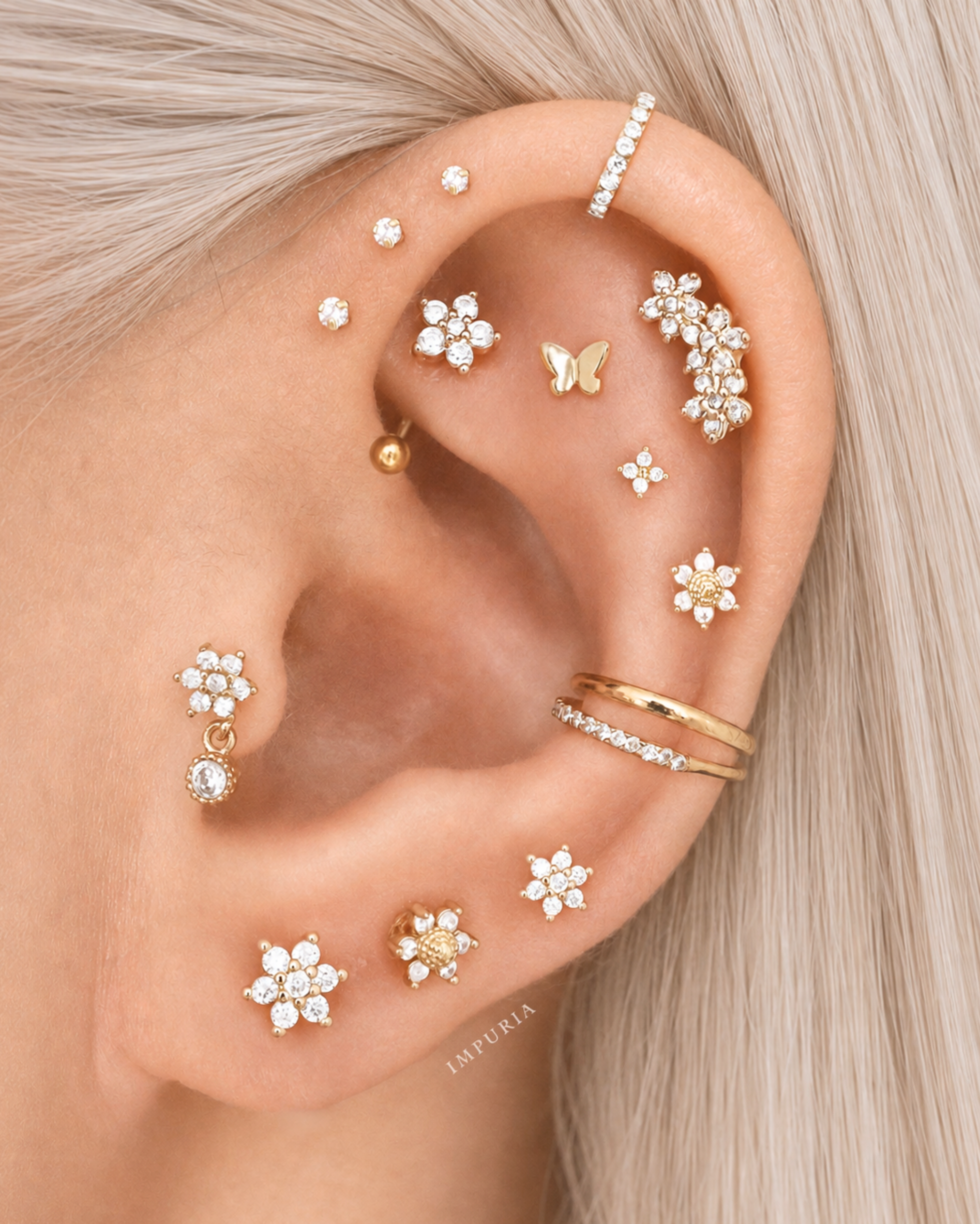 A close-up of a model's ear adorned with multiple gold and crystal stud earrings, floral cartilage piercings, a butterfly cartilage piercing, and an ear cuff, with blonde hair framing the ear.