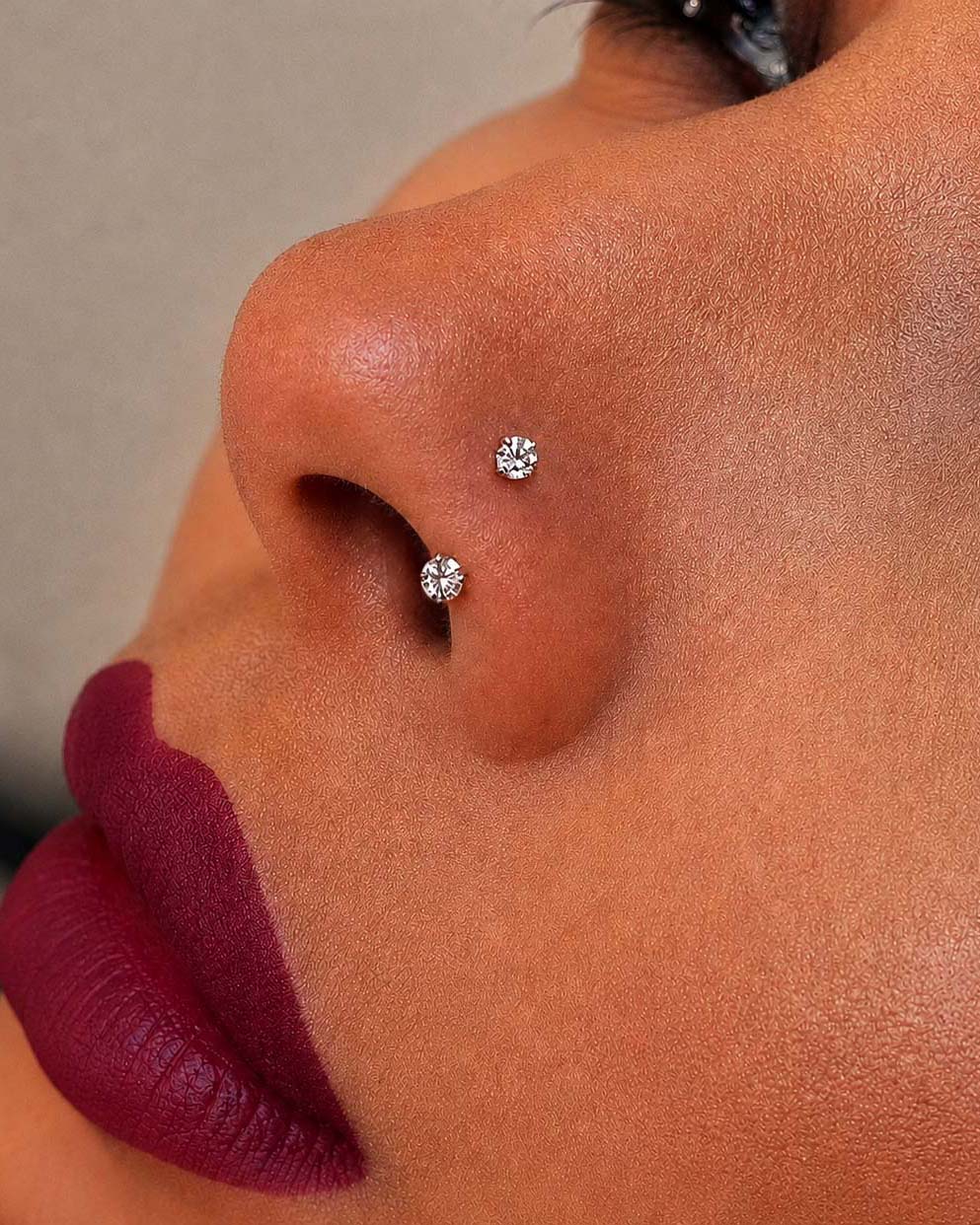 Close-up of a person's nose with two diamond stud earrings on a neutral background
