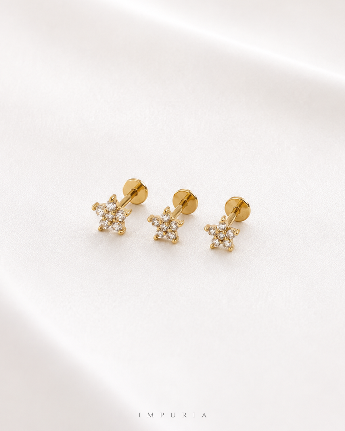 Three gold flower stud earrings with clear crystals, shown on a soft white fabric background with the brand name IMPURIA at the bottom.