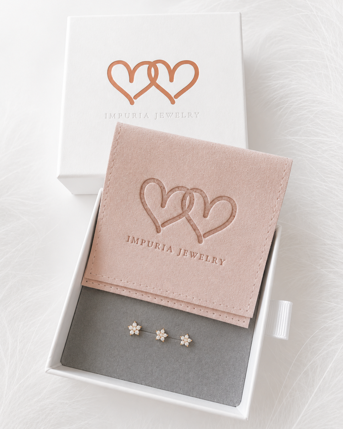Three small gold flower-shaped stud earrings displayed on a gray jewelry tray inside an open white gift box, with a blush pink velvet pouch and a white branded box lid behind it. The packaging is embossed with interlocking hearts and the brand name IMPURIA JEWELRY.