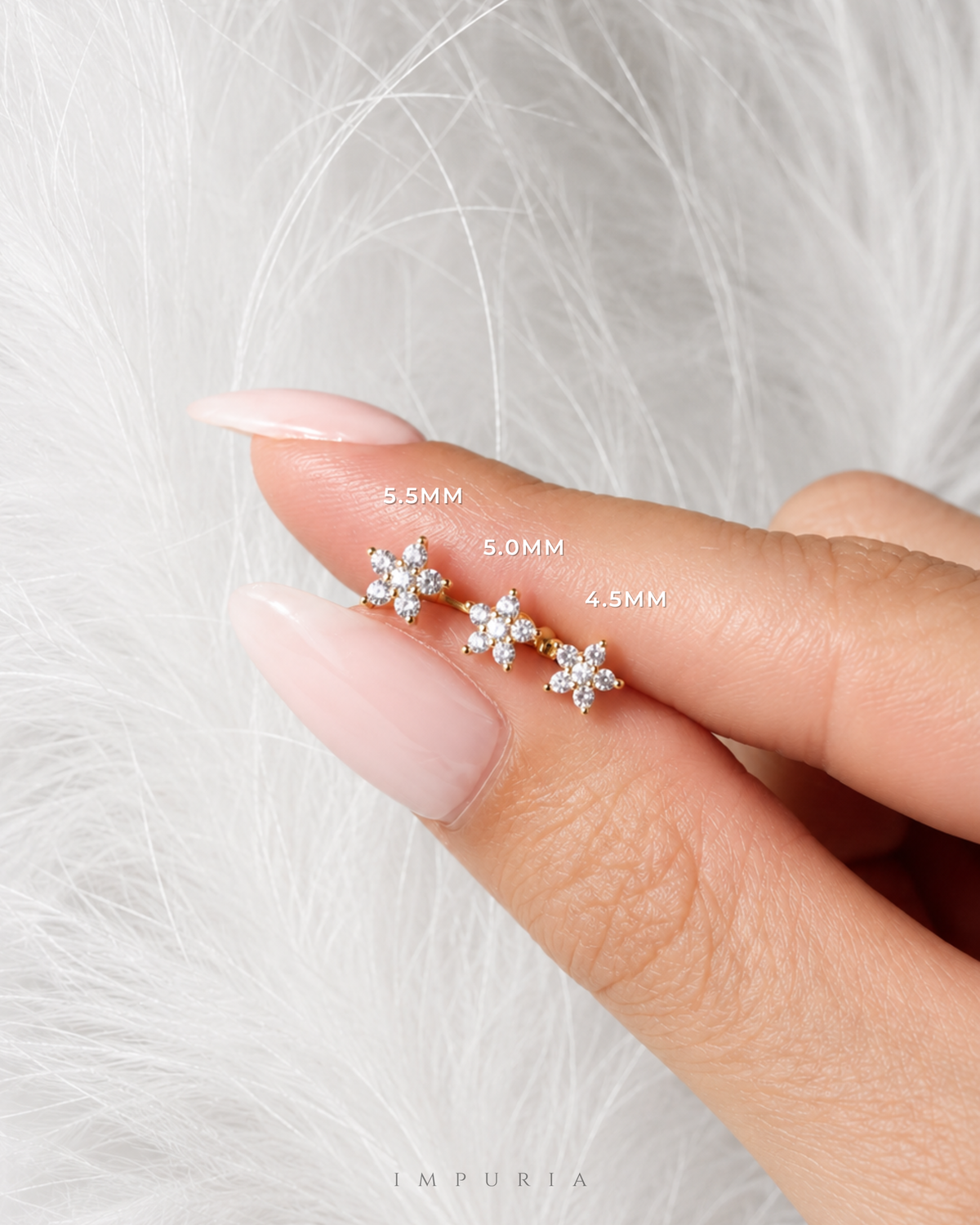A close-up of a hand with long nude manicured nails holding three small gold flower-shaped stud earrings with clear stones, labeled with sizes 5.5MM, 6.