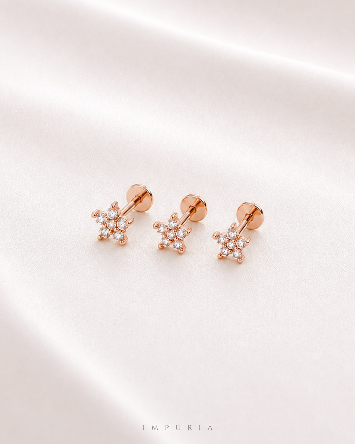 Three rose gold flower stud earrings with clear crystal details, arranged on a soft, draped white satin fabric background. The brand name 'IMPURIA' is subtly printed at the bottom center.