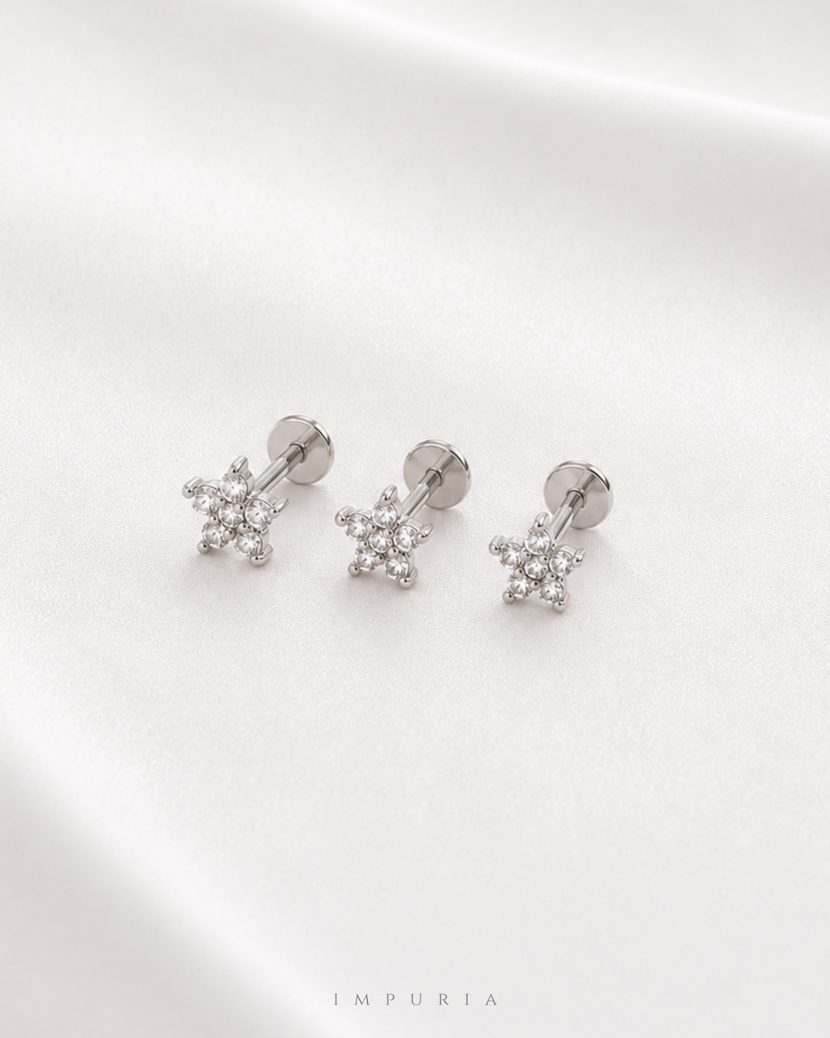 Three silver-toned flower stud earrings with clear crystal-like embellishments, arranged on a softly draped white fabric background. The brand name IMPURIA is subtly printed at the bottom center.