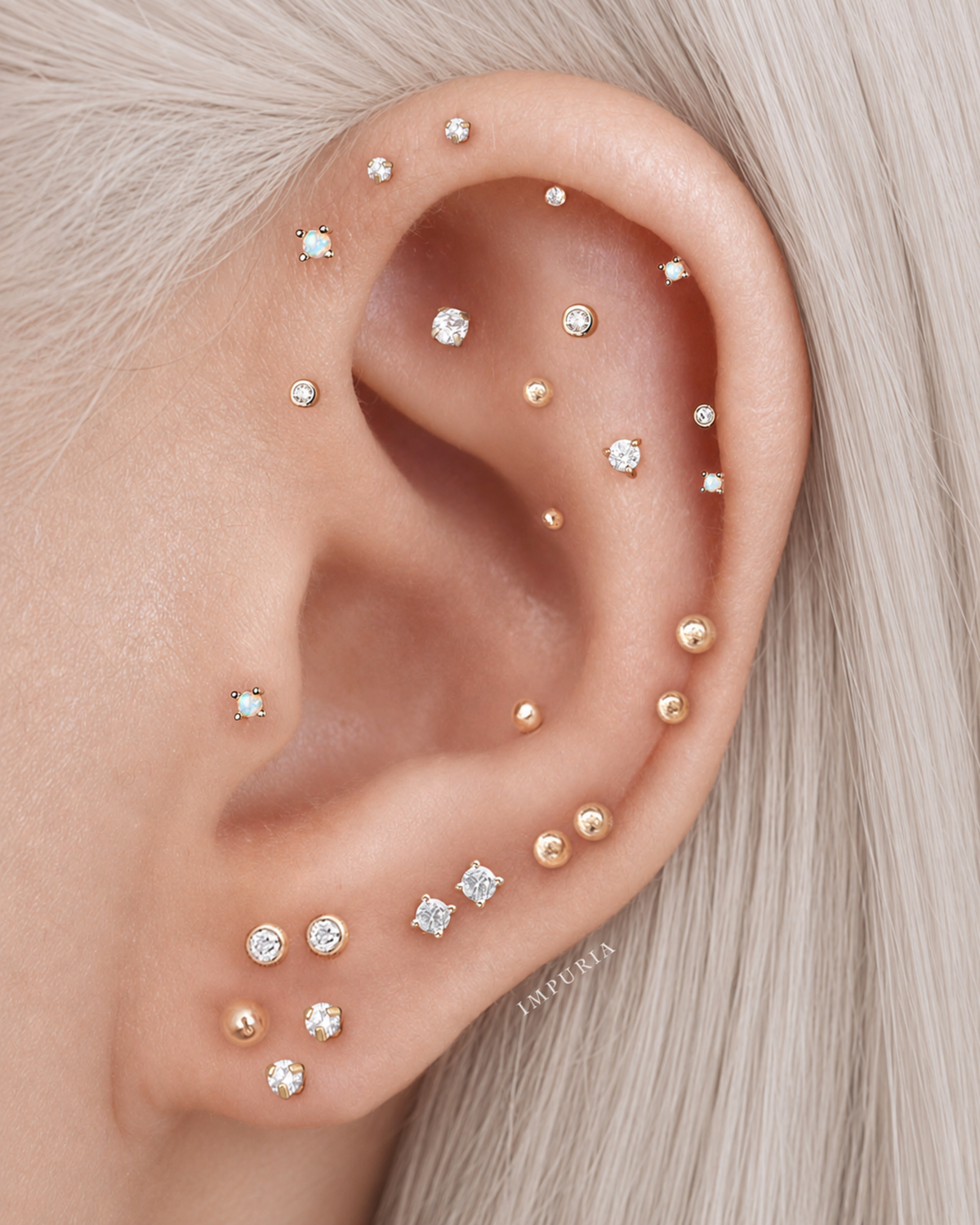Close-up of a human ear adorned with multiple small stud earrings in gold and clear gemstone settings, shown against pale blonde hair.